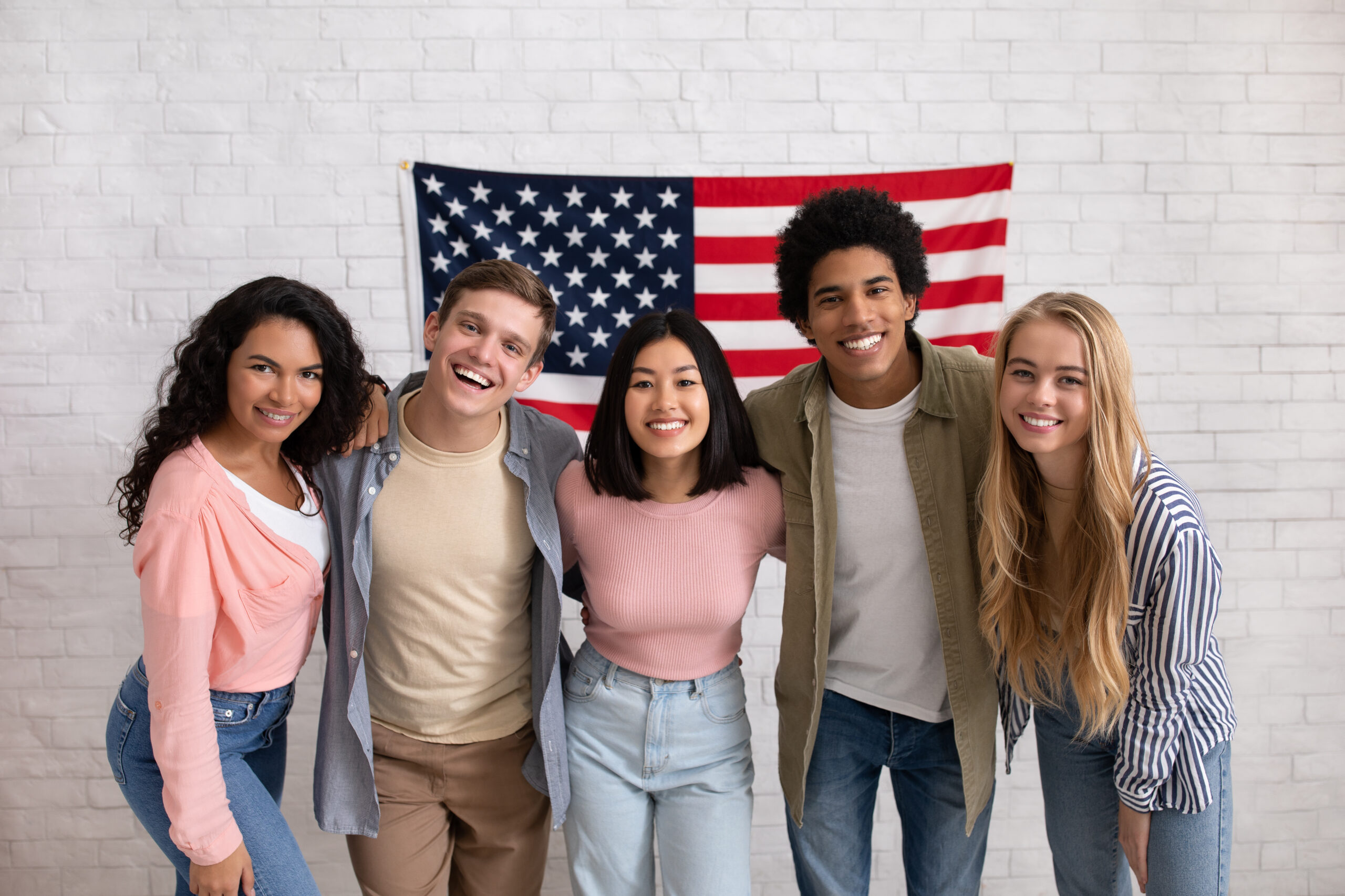 Fun education, language courses, learn english and exchange students. Young smiling people of different nationalities hug in college with large USA flag on white brick wall, free space, studio shot